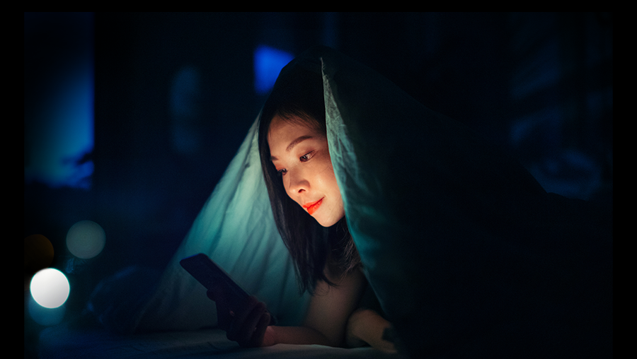 Asian woman looking at phone under bed covers
